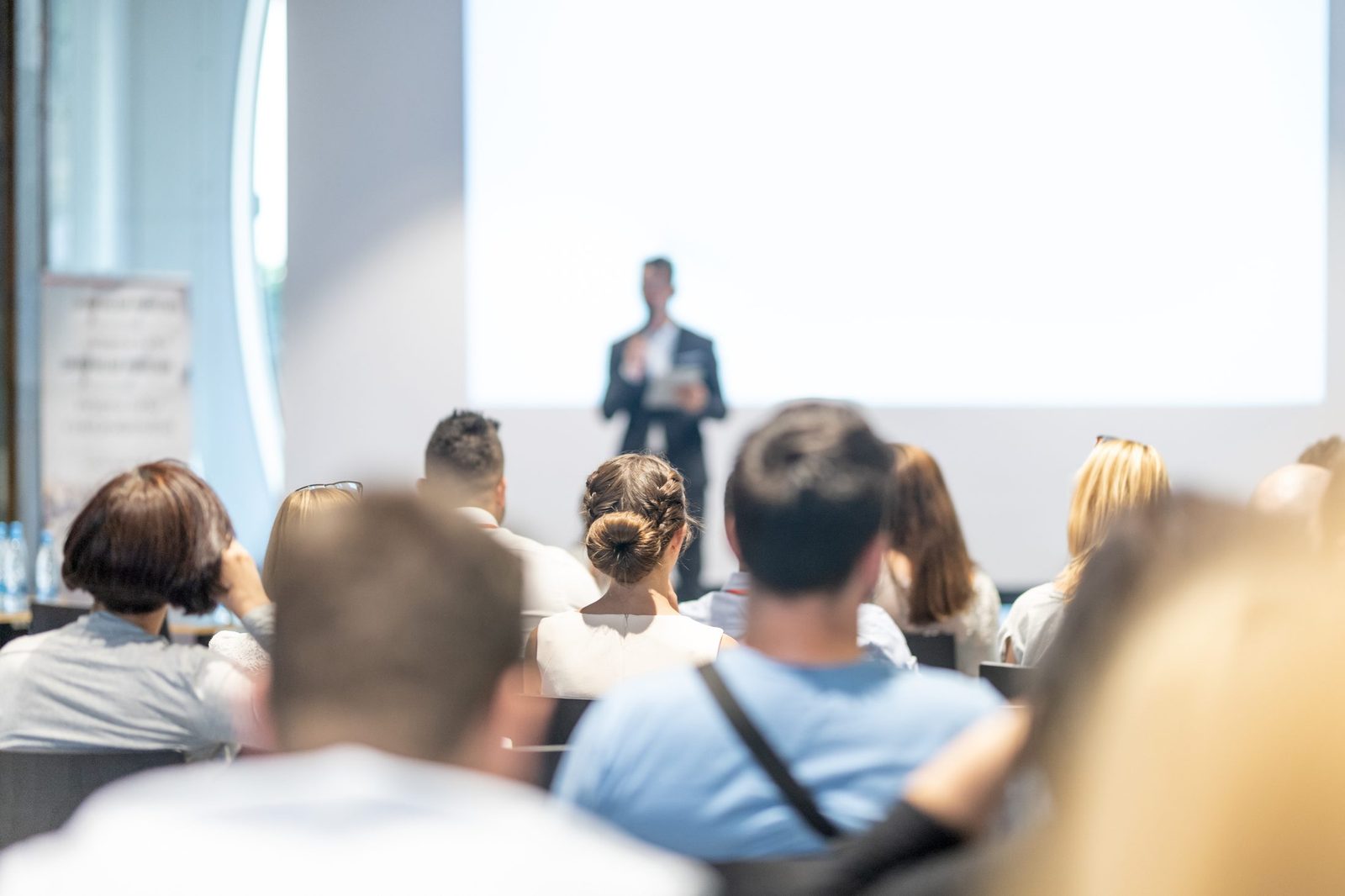 An audience listening to a presenter at the front of a bright room